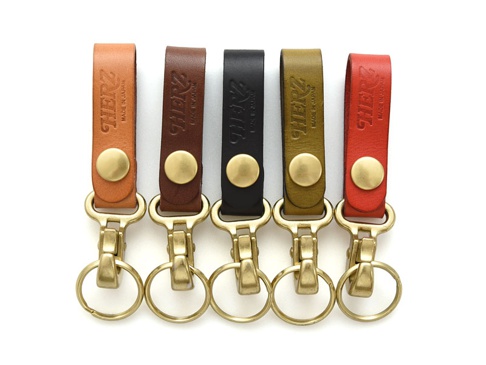 五個職人手作皮革鉤扣鑰匙圈，顏色從左至右分別為：淺棕色、深棕色、黑色、橄欖綠、紅色。皆為真皮材質，搭配黃銅色金屬鉤扣與鑰匙圈，皮革上壓印有「HERZ」品牌字樣。