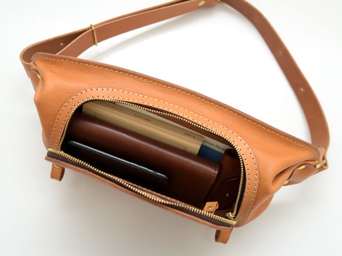 棕色皮革拉鍊旅行斜背包，主隔層打開，內有手機、皮夾和書籍。包包採用皮革材質，配有可調節的肩帶和黃銅色拉鍊。