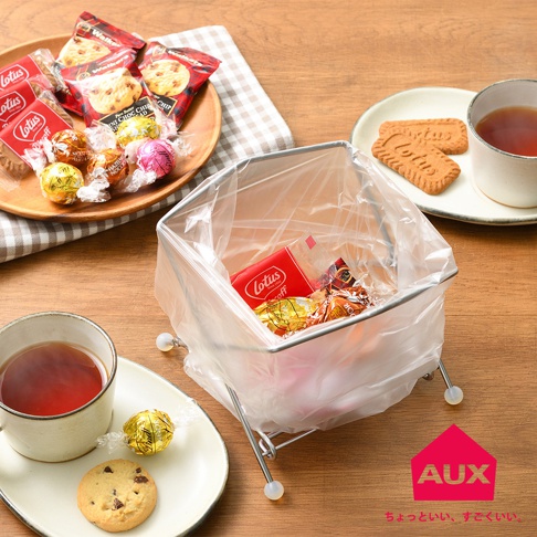 銀色金屬框架的折疊式垃圾架，內部裝有透明垃圾袋，裝有餅乾和巧克力。垃圾架的四個角落有白色圓形腳墊。背景是木製桌面，擺放著裝有餅乾和巧克力的木盤，以及兩杯茶。