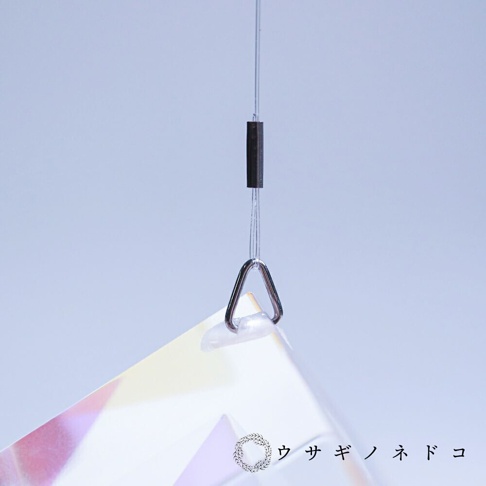 透明稜鏡立方塊吊飾，透過金屬環懸掛，底部可見部分彩虹色光澤，背景為淺藍色。