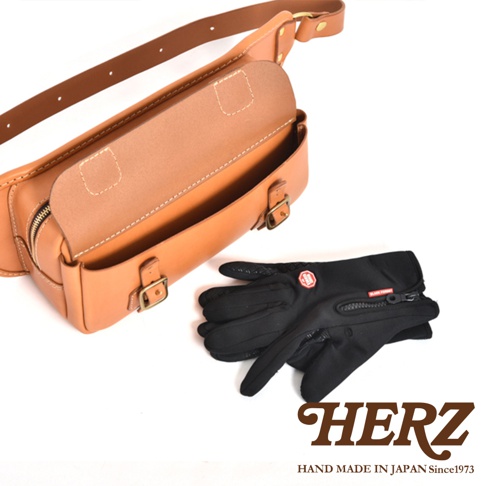 職人手作皮革騎士隨身斜背包，淺棕色皮革材質，設計簡約，附有可調節的皮帶和金屬扣環。一旁擺放一雙黑色騎士手套，手套表面有防滑紋路和拉鍊設計。