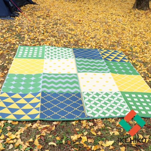 防水 PP 戶外野餐墊，以綠色、黃色、藍色和白色共四種顏色，拼貼多種幾何圖案，如條紋、圓點、菱形和方形，鋪設於秋日落葉的草地上。