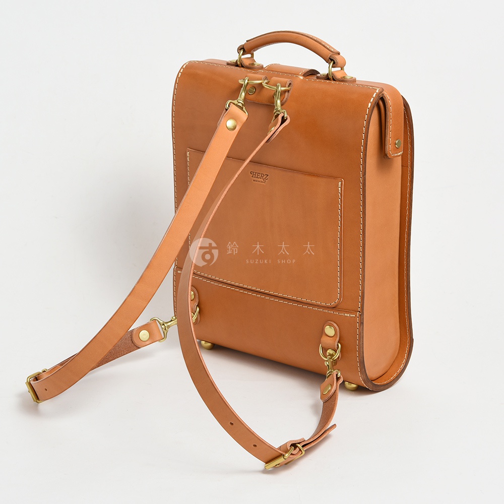 職人手作皮革杜勒斯商務後背包，採用淺棕色光滑皮革製成，配有可調節的兩條皮革背帶、頂部提把、黃銅色金屬扣環及底部支撐腳。包身線條簡潔，正面設有一個方形貼袋，印有品牌標誌。