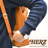 職人手作皮革後背包 R-143 加厚版，採用天然棕色皮革，搭配藍色紋理毛衣，展示側邊拉鍊與皮質背帶，手腕佩戴簡約腕錶。