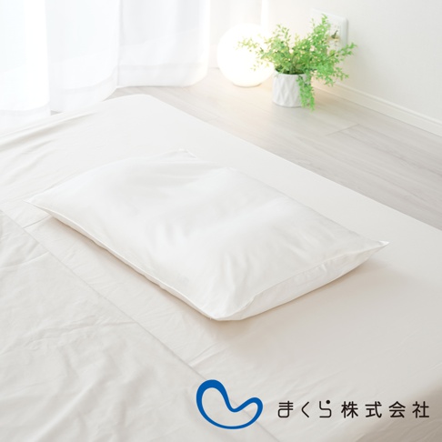 一張純白色枕頭套，由棉絲混紡材質製成，平整地放置在白色床單上，周圍環境明亮，有植物和燈光裝飾。