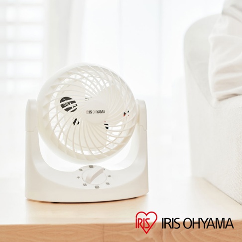白色IRIS OHYAMA桌上型空氣循環扇TAF-MKM10N，設有可調速的旋鈕，簡約的設計擺放在木質表面上，背景為柔和的白色窗簾與沙發。