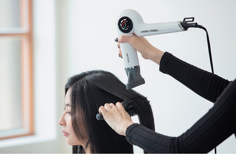一位女性正在使用 Tescom 離子吹風機為另一位女性吹整頭髮，吹風機為白色，帶有銀色邊飾和圓形溫度顯示。