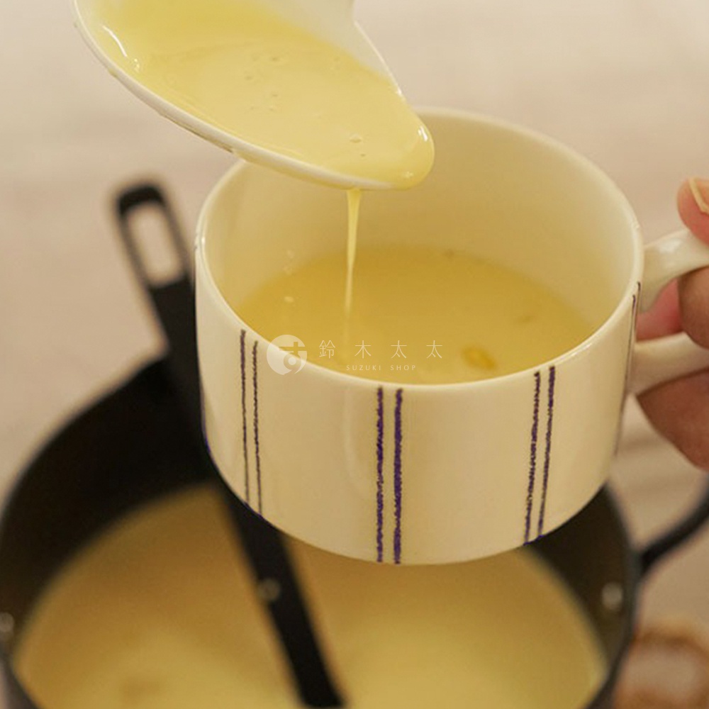 米色美濃燒手繪馬克杯，杯身飾有紫色線條，杯內盛有黃色液體，勺子正舀取液體。