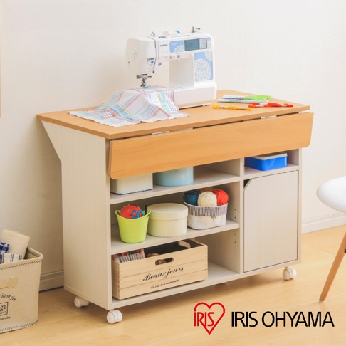 四層置物收納推車，上層為木紋桌面，桌面中央有可展開的木紋延伸板，上面擺放一台白色縫紉機、彩色布料及剪刀。推車主體為白色，有四層開放式層架及一個右側的白色門櫃，層架上擺放收納盒、毛線、木箱等物品，推車底部配有四個輪子。