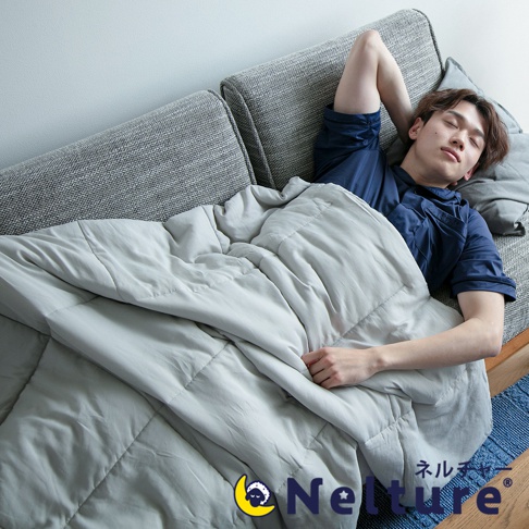 一件淺灰色、五層設計的單人涼感被，覆蓋在一名男子身上，他正躺在灰色沙發上休息。被子表面有菱格車縫線，材質看起來柔軟輕薄。