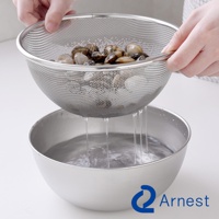 一只手正拿著一個不鏽鋼網篩，篩網裡裝滿了蛤蜊，水正從篩網中滴落到下方一個不鏽鋼盆裡。