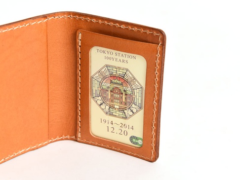 淺棕色皮革製成的三面雙折票券收納夾，內部有一個透明夾層，內含一張印有東京車站百年紀念圖案與日期的票券。