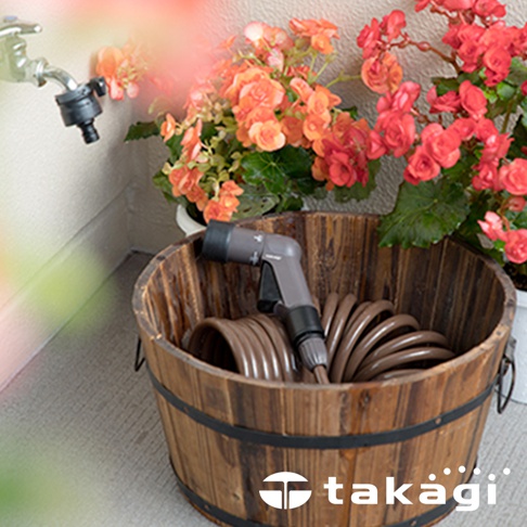 棕色螺旋狀的室外澆花水管，配有灰色噴嘴，收納在木桶中，背景有橘紅色和粉紅色花朵。