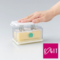 透明塑膠盒內裝有黃色洗衣皂，下方為白色底座與黃色刷毛。盒蓋為白色，內有兩根彈簧，一隻手正將盒蓋壓下。