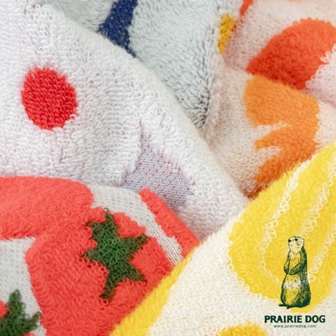 這是一張特寫圖片，展示了幾塊堆疊在一起的毛巾，毛巾材質柔軟，表面有提花刺繡圖案，圖案包含鮮豔的紅色圓點、橙色和黃色的葉子形狀，以及綠色的星星和紅色的番茄圖案。圖片右下角有一個草原犬鼠的插畫，下方有PRAIRIE DOG和網站地址。