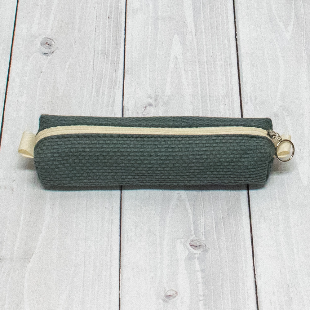 深綠色三河木綿刺子織長條形筆袋，配米白色拉鍊和黃色提帶，拉鍊頭為銀色圓環。