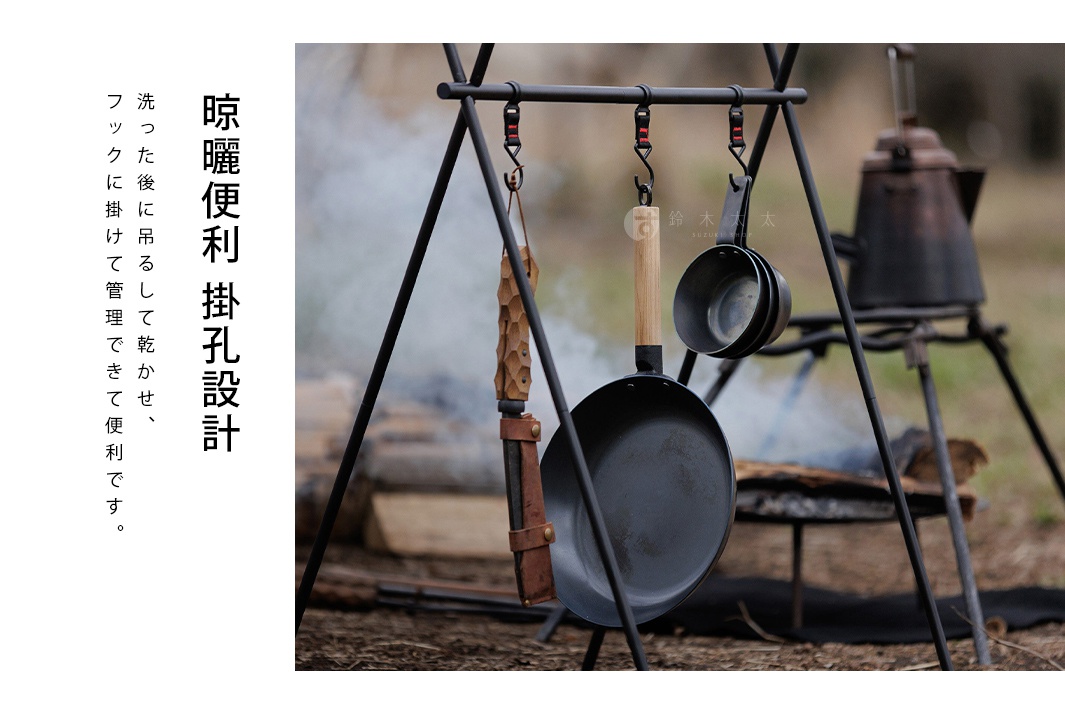 黑色鐵鍋、木柄，以及兩個尺寸不同的銀色鐵杯，懸掛在一個黑色三腳架上，背景是戶外營火及煙霧。