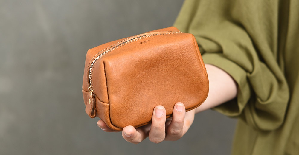 一位穿著橄欖綠色衣服的人手持一個暖棕色皮革收納包，包身為長方體結構並搭配金屬拉鍊，皮革表面呈現自然的紋理質感，包身角落處有細緻的 Organ 品牌壓印，整體呈現簡約的職人手作風格。