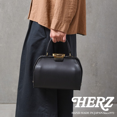 黑色皮革手提包，設有金色金屬扣和提把，包身線條硬挺，風格復古。