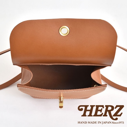 職人手作皮革翻蓋迷你後背包，採用棕褐色真皮製作，包身結構紮實，翻蓋處有金色圓形金屬扣眼，包口處為金色鎖扣，細緻的白色縫線展現手工質感。