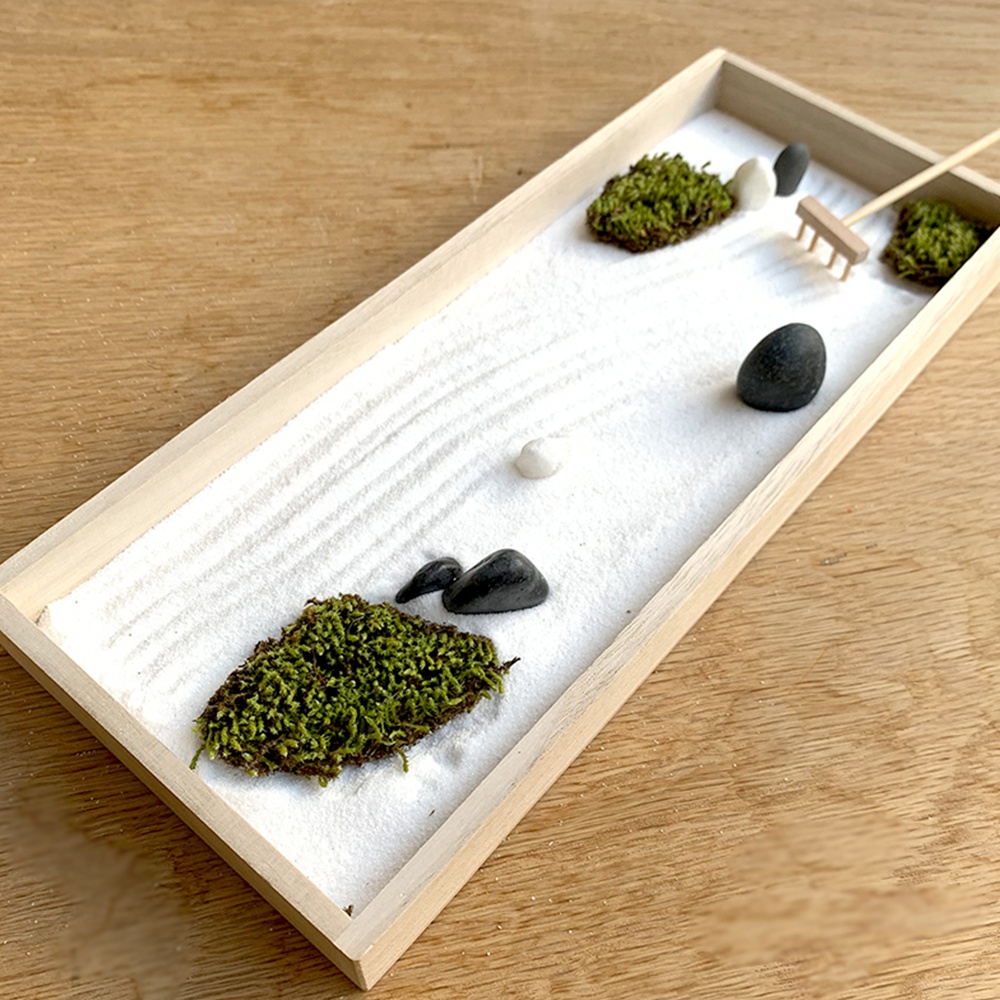 淺木色長方形托盤內，鋪著細膩的白色沙子，上面有耙出的波紋。沙中點綴著幾簇翠綠色的苔蘚、光滑的黑色鵝卵石和白色小石子。右側有一把迷你木製耙子。