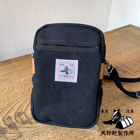 黑色石洗帆布斜背手機包，附有白色方形標籤，上面印有日文圖案。包包設有拉鍊開口及肩帶。