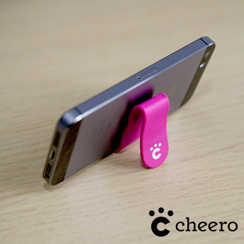 一個桃紅色的萬能QQ變形夾，由柔軟的矽膠材質製成，夾住一支深灰色的手機，手機平放在木紋桌面上。變形夾上印有白色的品牌標誌，是一個帶有爪印的字母「C」。