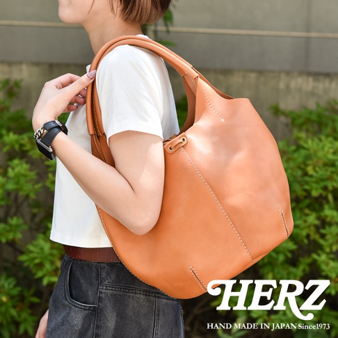 一位女性肩掛著一個棕色皮革球形托特包，包包採用縫線裝飾，搭配黑色牛仔褲和白色上衣。