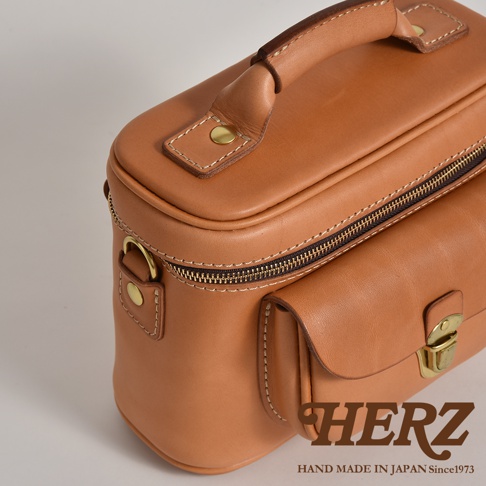 職人手作皮革相機包，淺棕色軟皮材質，帶有明顯的縫線細節、黃銅色拉鍊、金屬扣件與提把，包身側面有小口袋設計。