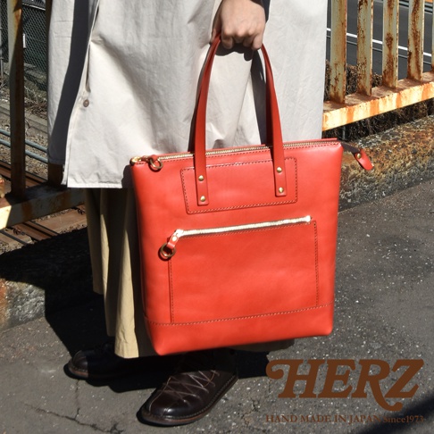 一位模特手提著日本HERZ品牌的紅色皮革托特包，該包款呈現簡約的直式設計，包身正面設有金色拉鍊口袋，整體散發精緻的職人手工質感，背景為戶外街道場景。