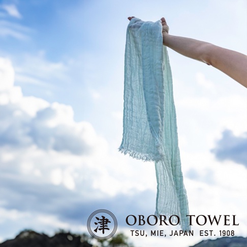 一隻手高舉著一條淺藍色的三層紗純棉掛脖毛巾，毛巾上有細微的皺褶和流蘇邊緣，背景是晴朗的天空和白色雲朵。