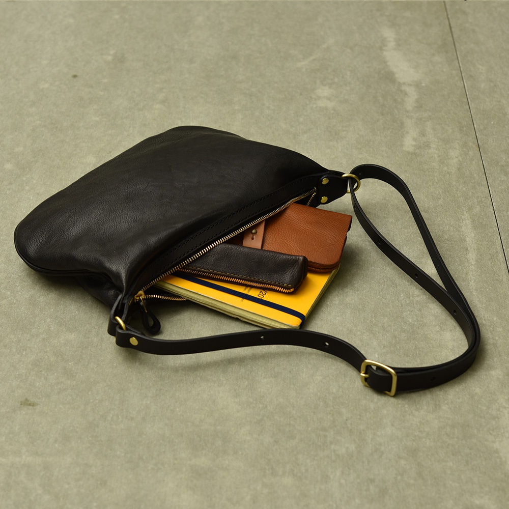 黑色皮革職人手作Organ單肩半月包，包內裝有黃色筆記本、棕色皮夾及黑色小皮件。包身線條簡潔，搭配金屬拉鍊與可調式單肩背帶。