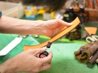 職人手作皮革筆筒製作過程，雙手正使用打磨工具處理淺棕色皮革邊緣，背景為工作檯與其他工具。