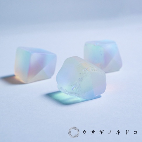 三顆半透明的合光稜鏡石，呈現柔和的彩虹光澤，有立方體和不規則多面體的形狀，材質看似玻璃或水晶，散發著淡藍、粉紅和淡黃色的光芒。