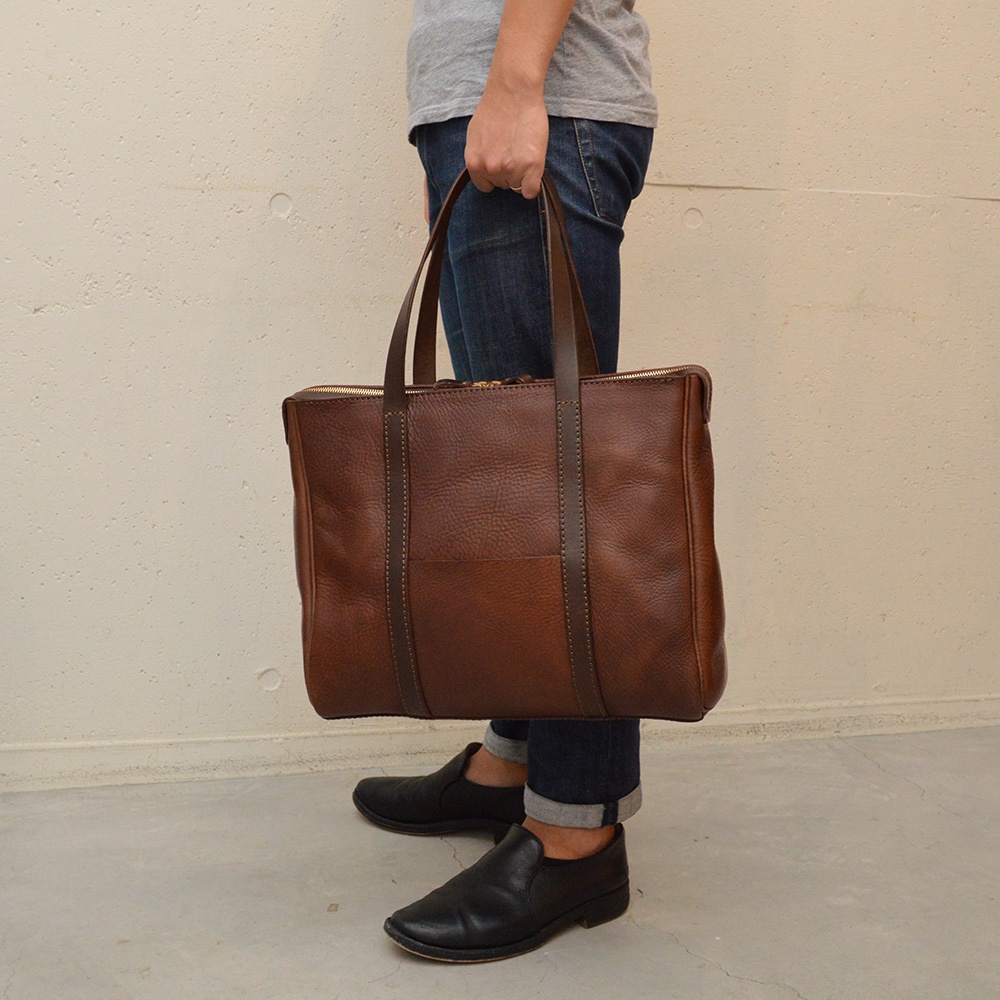 棕色皮革製成的手提肩背包，帶有拉鍊開合和兩條提把，包包正面有一個嵌有皮革的口袋。