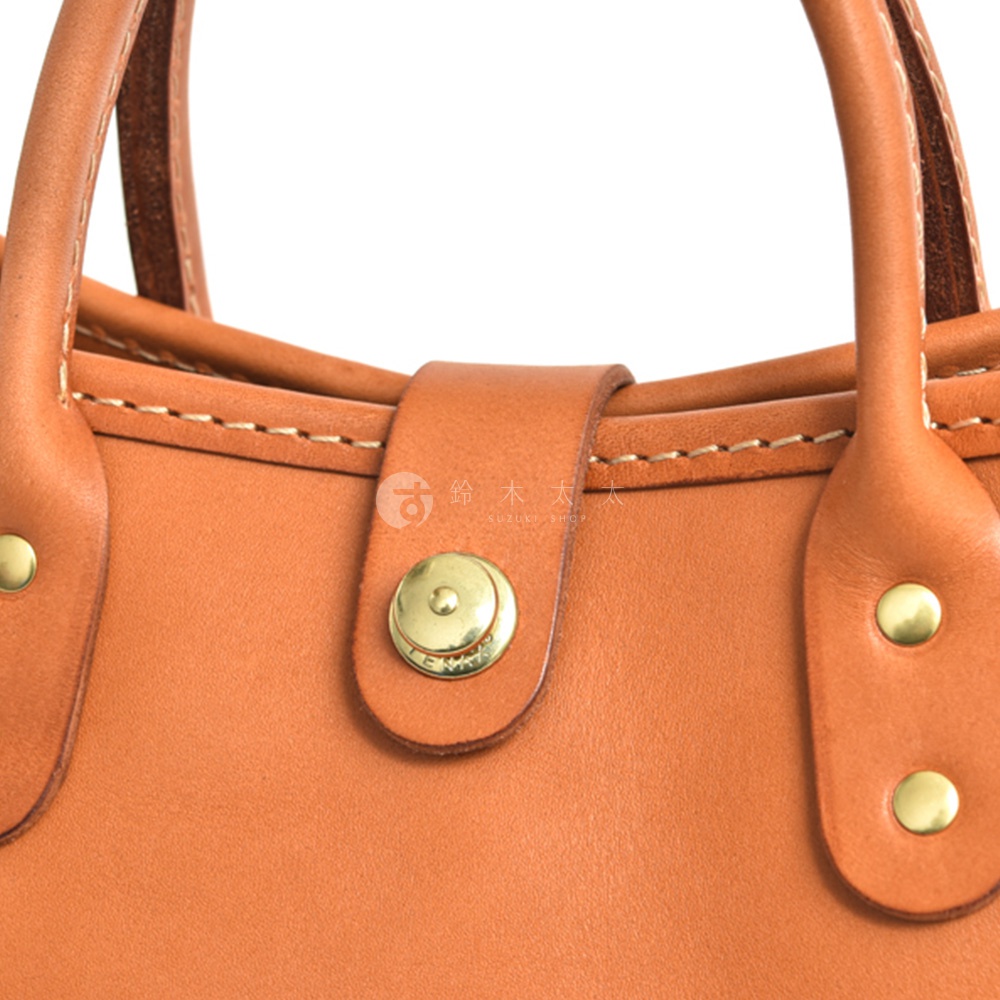 職人手作皮革迷你托特包，呈現溫潤的橘棕色調，包身以細緻的縫線勾勒，搭配品牌壓紋的黃銅旋鈕釦和鉚釘，展現皮革的質感與工藝。