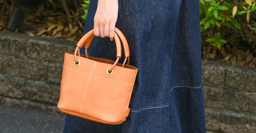 橘色皮革製成的午餐袋，配有加厚的皮革提把，袋口寬大，有縫線裝飾，並有金色金屬孔眼。