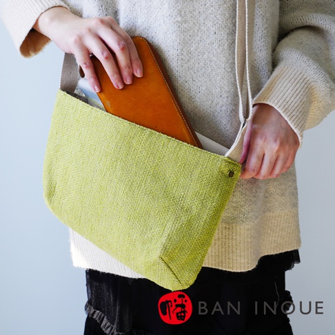 BAN INOUE 品牌字樣出現在圖片右下角，左側有一女性穿著淺灰色毛衣，雙手正將一個棕色長夾放入綠色織紋手提袋中。