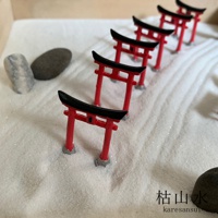 一排紅黑色鳥居模型，立於細白沙地上，旁邊散落著幾顆灰黑色石塊。