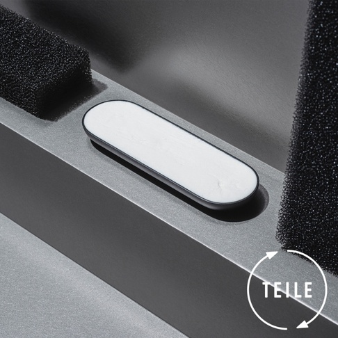 一個橢圓形的白色固體皂，帶有黑色邊框，放置在一個傾斜的灰色平台上。平台旁邊是兩個黑色的粗糙海綿塊。