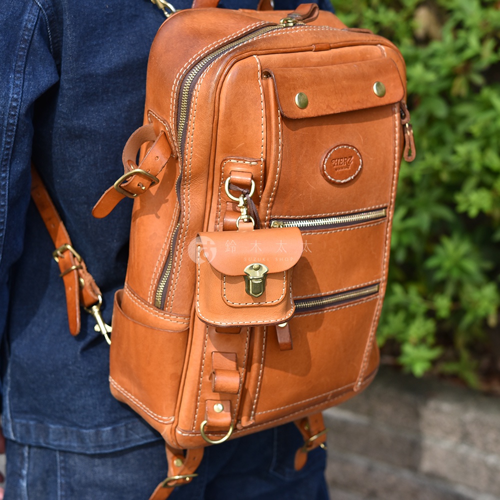 一款棕色皮革製成的耳機收納包，搭配金色金屬扣環，懸掛於同色系皮革背包上。