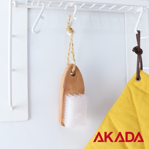 赤田 AKADA 品牌標誌下，懸掛著一個木質手柄的白色纖維刷，旁側掛著一個黃色菱格紋布料。