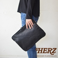 手持一個黑色職人手作皮革Organ手拿包，包體呈現柔軟的皮革質感，搭配牛仔褲與黑色上衣。