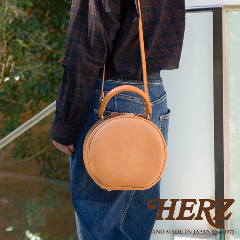 HERZ 品牌標誌與 HAND MADE IN JAPAN Since1973 字樣，下方展示一款棕褐色皮革製成的圓形側背包。