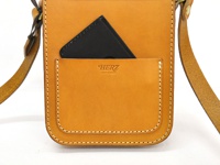 職人手作皮革皮扣直式單肩包，採用鞣製皮革製成，呈現溫潤的焦糖棕色。包款正面有一個大口袋，內嵌一個黑色手機。包包的邊緣以白色車線縫製，質感樸實。