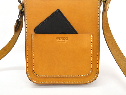 職人手作皮革皮扣直式單肩包，採用鞣製皮革製成，呈現溫潤的焦糖棕色。包款正面有一個大口袋，內嵌一個黑色手機。包包的邊緣以白色車線縫製，質感樸實。