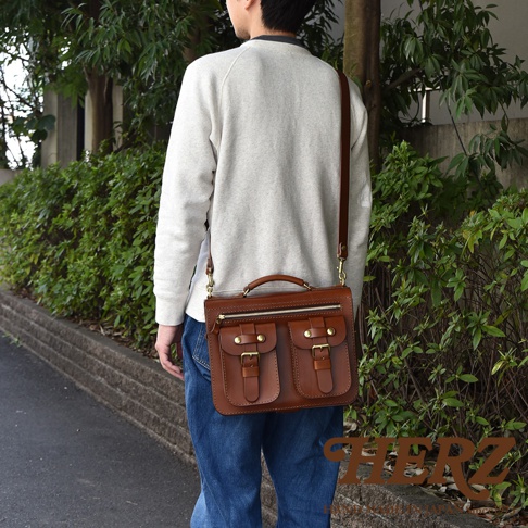 職人手作皮革雙口袋兩用單肩包，採用棕色皮革製成，設有兩個前置翻蓋口袋，以皮帶和金屬扣固定，頂部配有手提把，可斜背或側背。