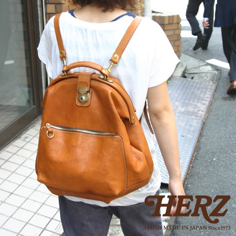 職人手作皮革輕旅行後背包 R-144，呈現溫暖的焦糖棕色。包款採用柔軟的皮革材質，配有金屬扣環的頂部提把和可調式肩帶，正面有一個拉鍊口袋和一個貼袋，整體設計簡潔實用。