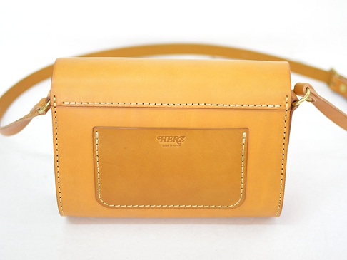 職人手作皮革寬扣帶馬鞍包，以天然皮革製成，呈現溫潤的焦糖色。包款設計簡約，正面有一個方形外袋，邊緣有精緻的縫線裝飾。寬版的皮革背帶透過金屬環連接包身。