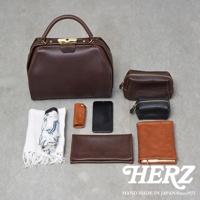 職人手作皮革Organ兩用口金手提包，深棕色皮革，配有金色金屬扣。旁邊擺放著一個深棕色化妝包、一個黑色化妝包、一個棕色皮革鑰匙包、一個黑色手機、一個對折的棕色皮革長夾、一條白色毛巾和一把摺疊傘。